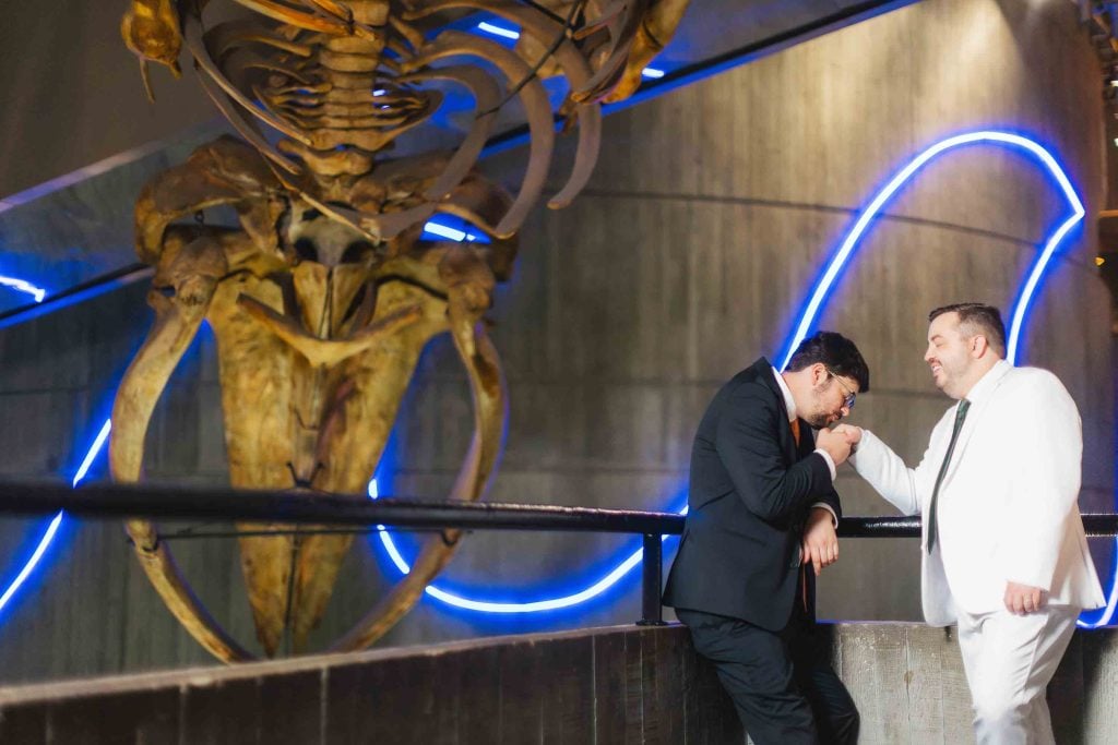 Two men in suits share a tender moment in front of a large whale skeleton display with blue neon lights, creating striking wedding portraits in a modern Baltimore museum setting.