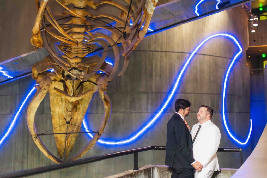 Two men in formal attire hold hands and smile at each other under a large whale skeleton display with blue neon lights on a concrete wall—capturing a unique Baltimore wedding moment.