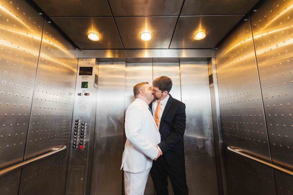 Two men in formal suits share a kiss while standing inside a brightly lit, stainless steel elevator, capturing wedding portraits.