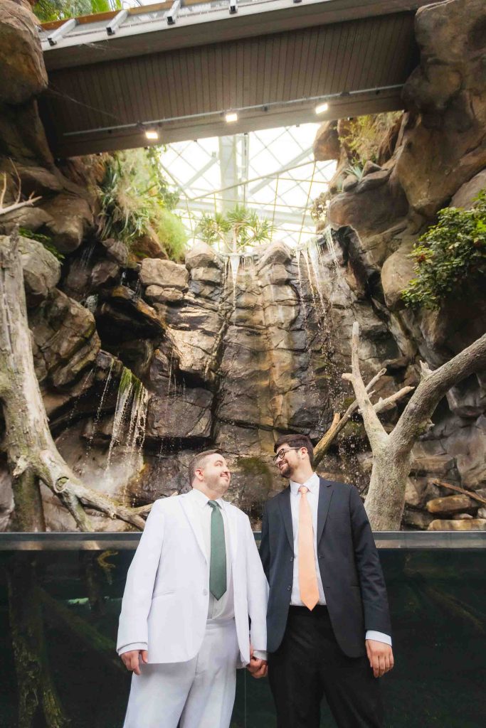 Two men wearing suits hold hands and look at each other, standing in front of the National Aquarium's indoor waterfall with rocks and greenery—a beautiful setting for wedding portraits.
