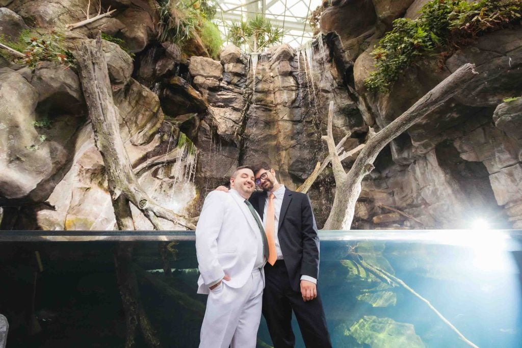 Two men stand close together in front of a large indoor rock waterfall and aquarium at the National Aquarium in Baltimore, both dressed in suits—one white, one black—with lush greenery and shimmering water behind them.