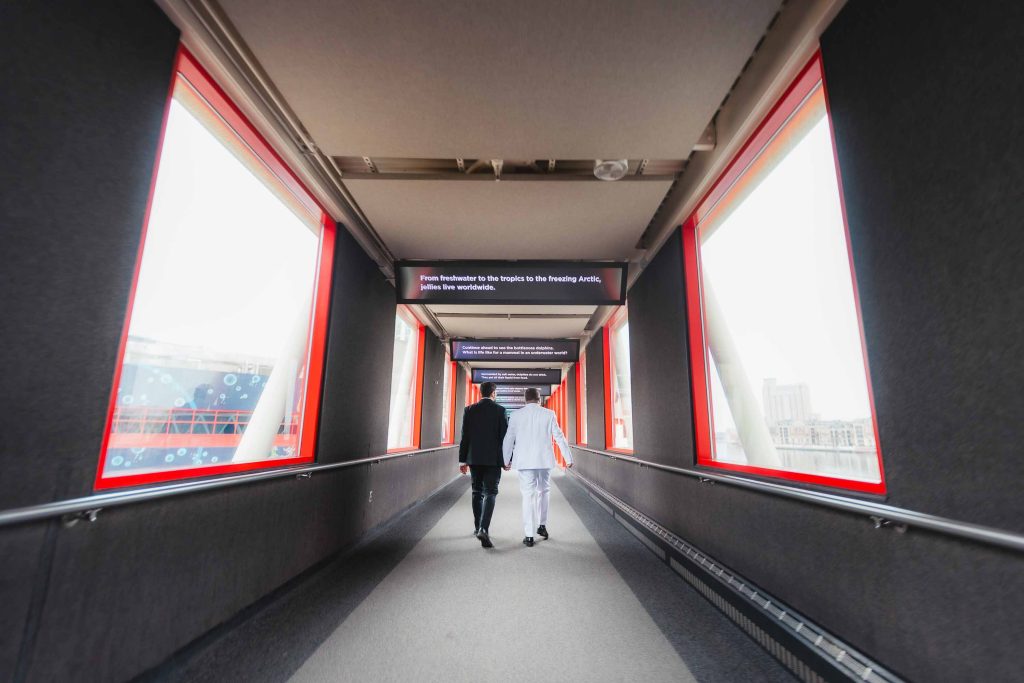 Two people walk down an enclosed hallway with large windows and red frames at the National Aquarium in Baltimore, heading toward a sign with directions.