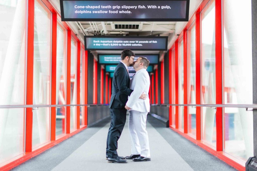 Two men, dressed in black and white suits, share a kiss in a brightly lit walkway with red pillars and informational signs above—capturing wedding portraits filled with love and joy.