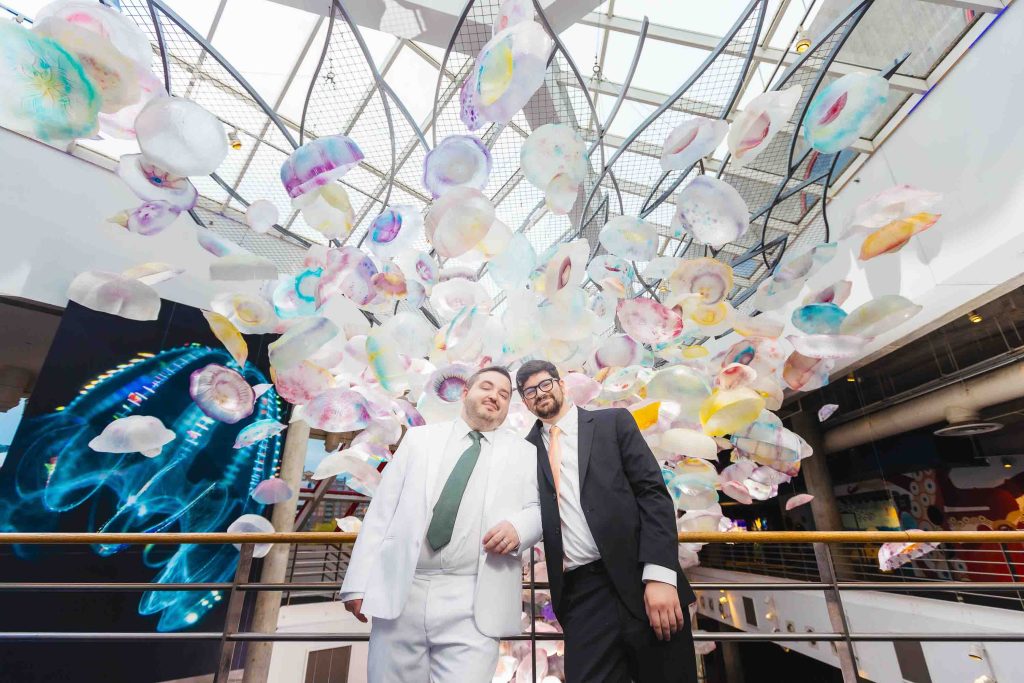 Two people in suits stand together indoors beneath an art installation of suspended, colorful jellyfish-like sculptures under a glass ceiling—capturing unique wedding portraits in the heart of Baltimore.