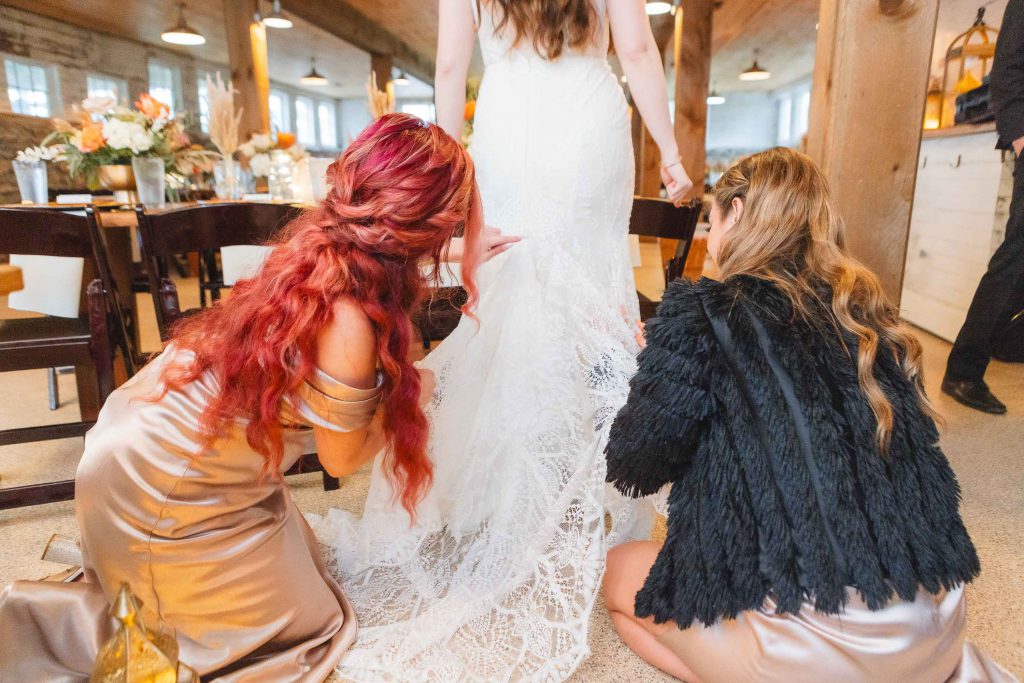Two women in formal dresses adjust the lace train of a bride's white wedding gown inside The Barns at Hamilton Station, setting the perfect scene for an unforgettable wedding reception.