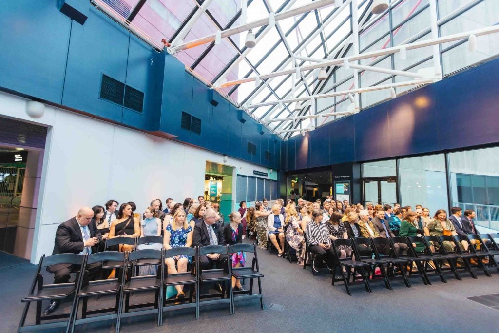A group of people seated in rows of chairs in a modern, glass-roofed atrium at the National Aquarium, facing forward as if waiting for a ceremony to begin.