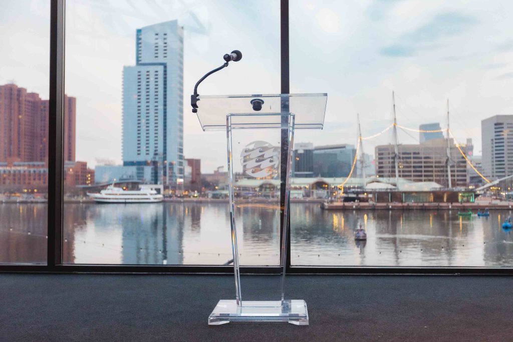 A clear podium with a microphone stands in front of large windows overlooking Baltimore’s city skyline and waterfront—perfect for sharing wedding details or heartfelt speeches.