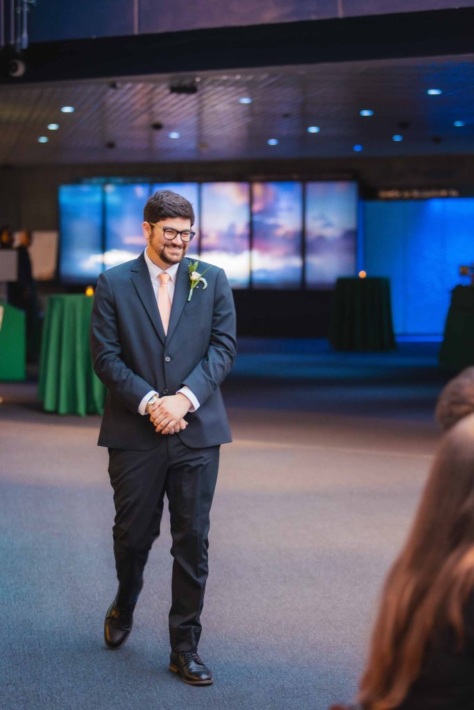 A man in a suit with a boutonniere walks indoors, smiling with hands clasped, under soft lighting at a wedding ceremony, with green-covered tables in the background.