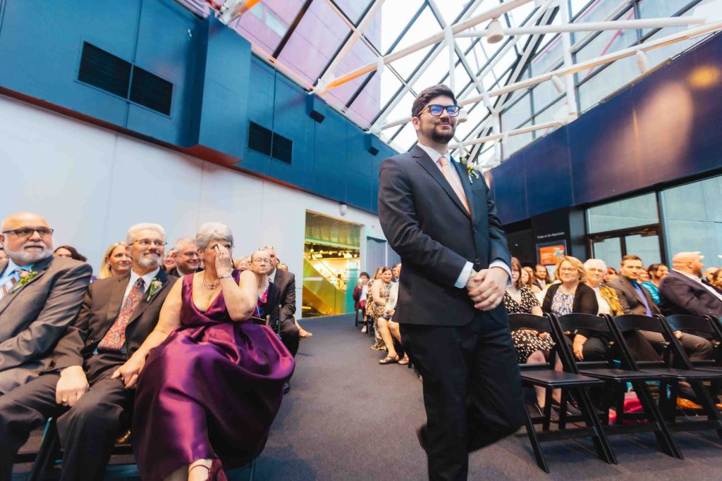 A man in a suit walks down the aisle at a same sex wedding, while seated guests watch and smile in a modern, glass-roofed venue reminiscent of the National Aquarium.