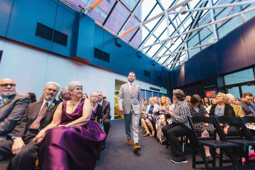 A man in a light gray suit walks down the aisle between seated guests at a same sex ceremony held indoors beneath a modern glass ceiling in Baltimore.