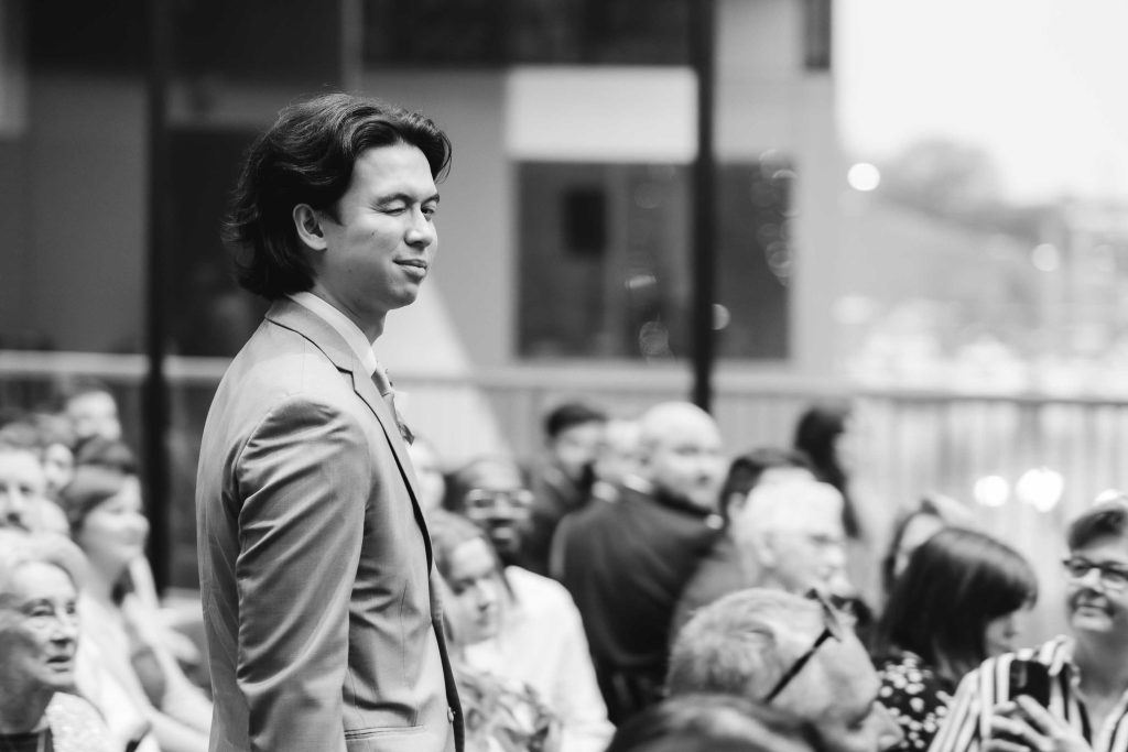 A man in a suit stands among a seated crowd at a same-sex ceremony, eyes closed and smiling slightly, in a black-and-white indoor setting.