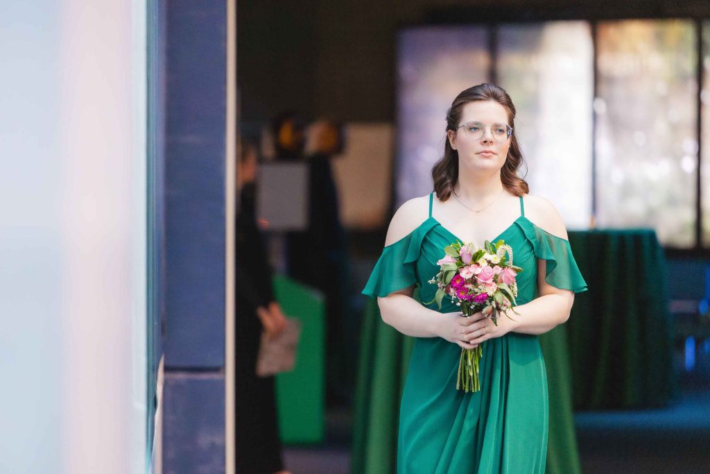 A woman in a green dress holding a bouquet of flowers stands indoors at a ceremony in Baltimore, looking ahead with a neutral expression.