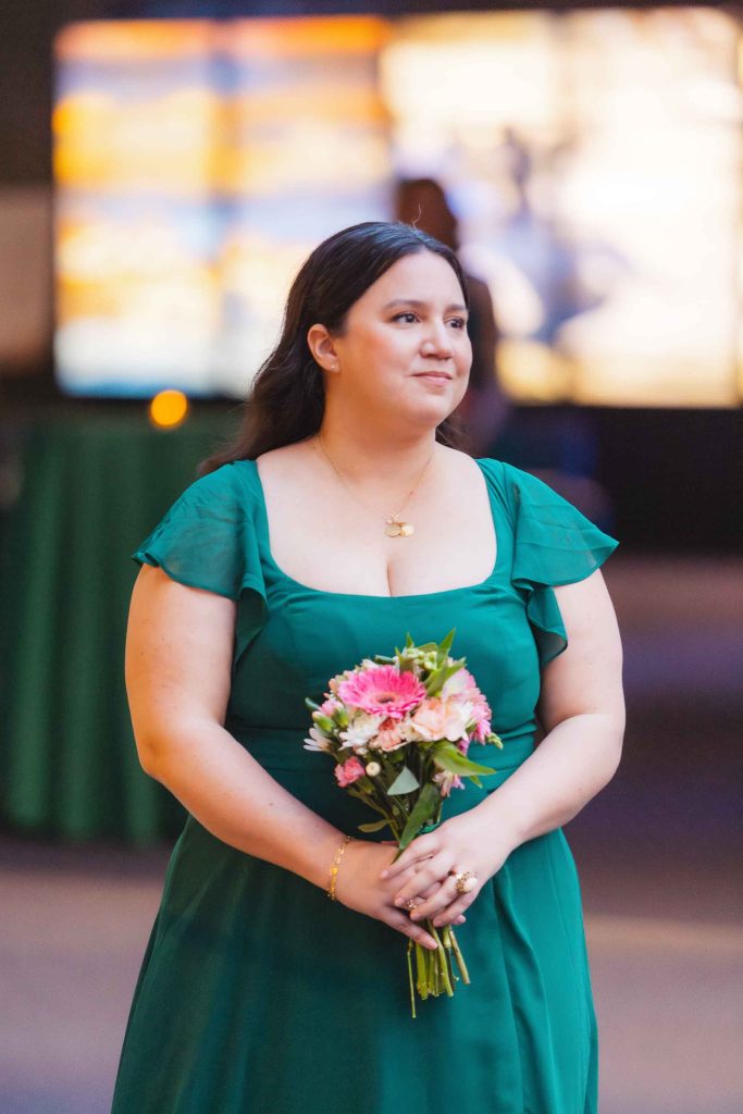 A woman in a green dress holds a bouquet of pink and white flowers, standing indoors at a same sex ceremony in Baltimore with blurred lights in the background.