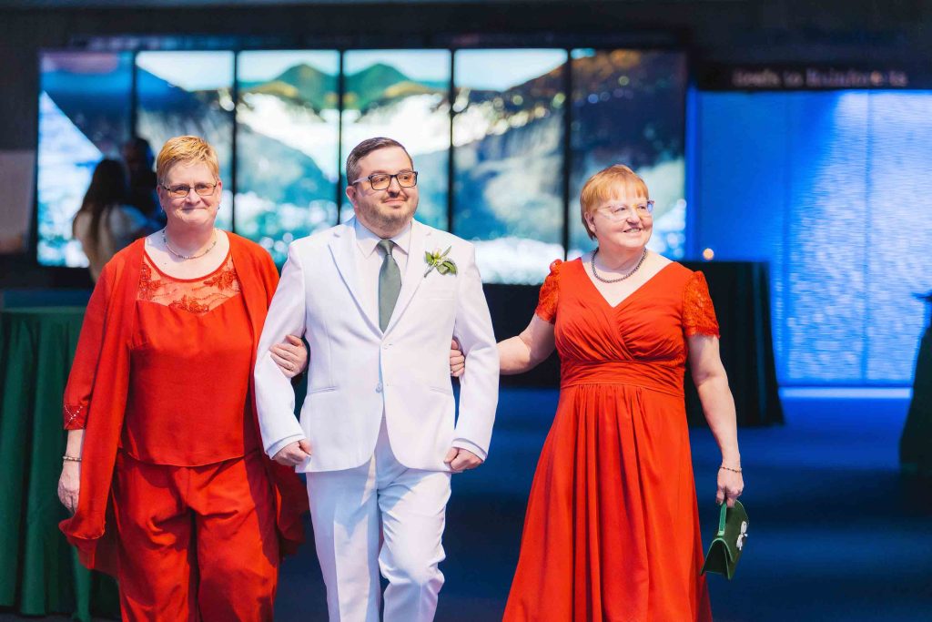 A man in a white suit walks arm in arm with two women in red dresses indoors at the National Aquarium, with a landscape image displayed on monitors in the background.