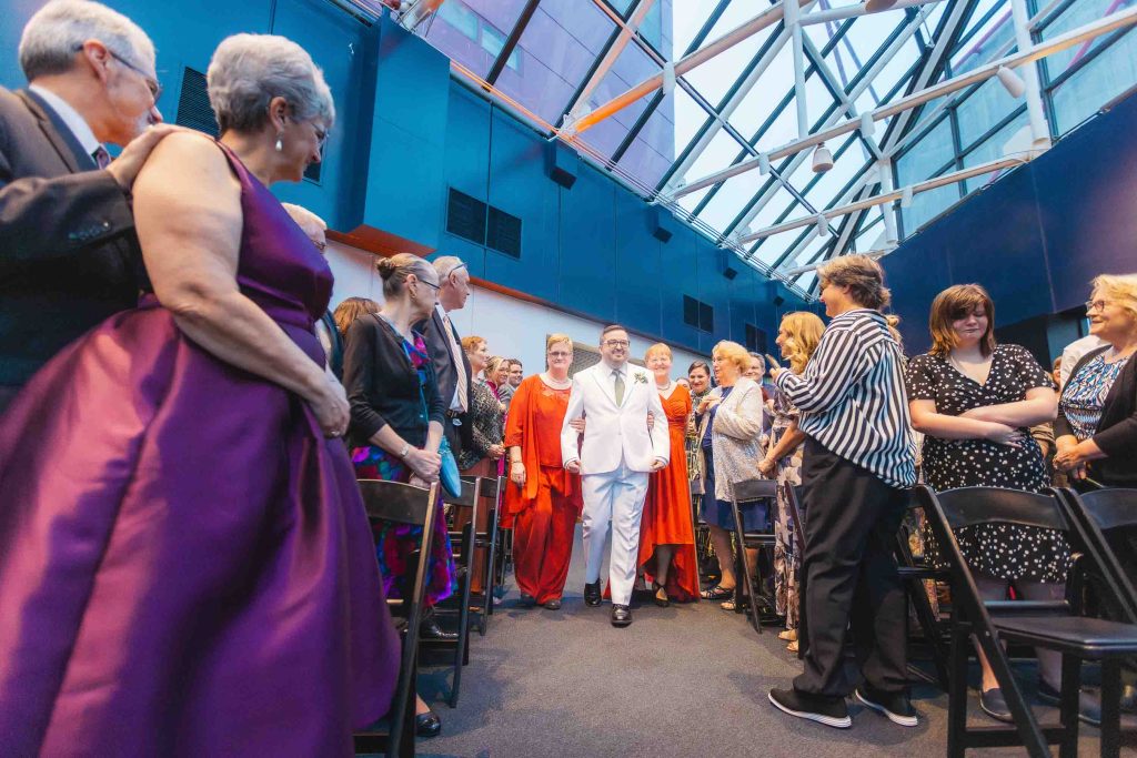 A man in a white suit walks down an aisle between seated and standing guests in formal attire at a stunning, glass-ceilinged Baltimore wedding venue.