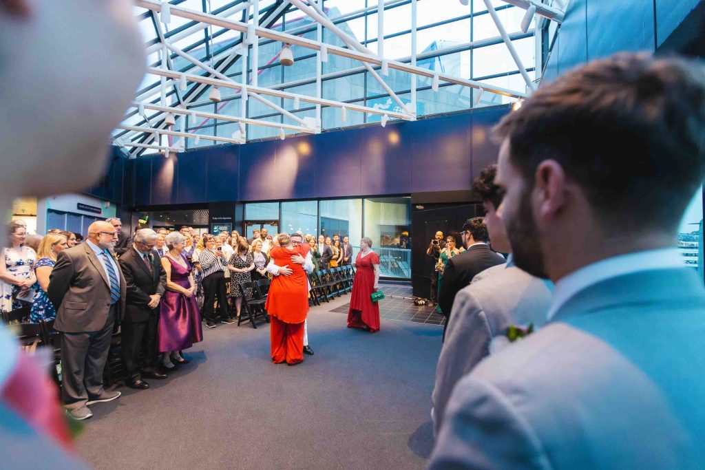 Two people in red embrace in the center of a modern indoor venue at the National Aquarium in Baltimore as guests, some standing and some seated, watch during a formal wedding event.
