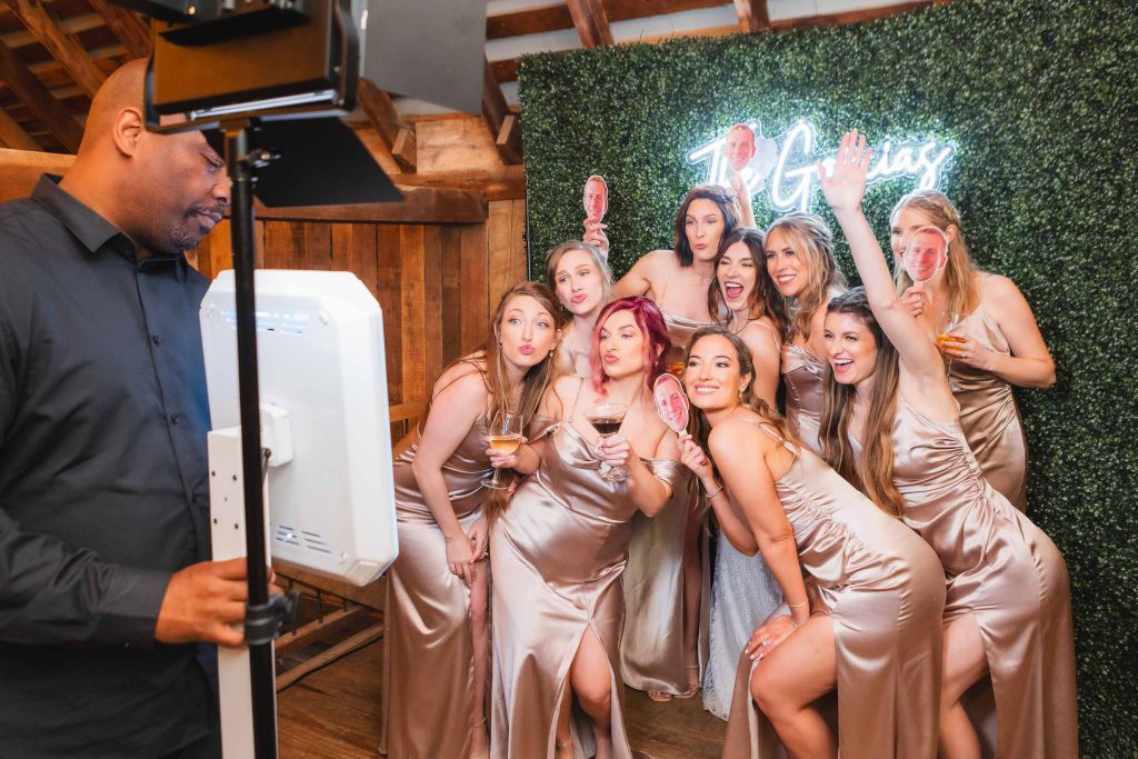 A group of women in light-colored dresses pose for a photo with an attendant operating a photobooth at a wedding reception at The Barns at Hamilton Station, featuring a greenery backdrop and neon sign.