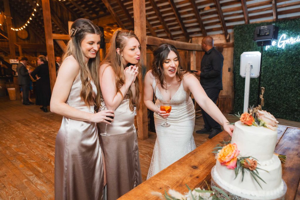 Three women in formal dresses stand by a decorated table with a tiered cake at The Barns at Hamilton Station. One woman in white reaches toward the cake, while the other two look on, one holding a drink. The charming wood-paneled room adds to the elegant wedding reception atmosphere.