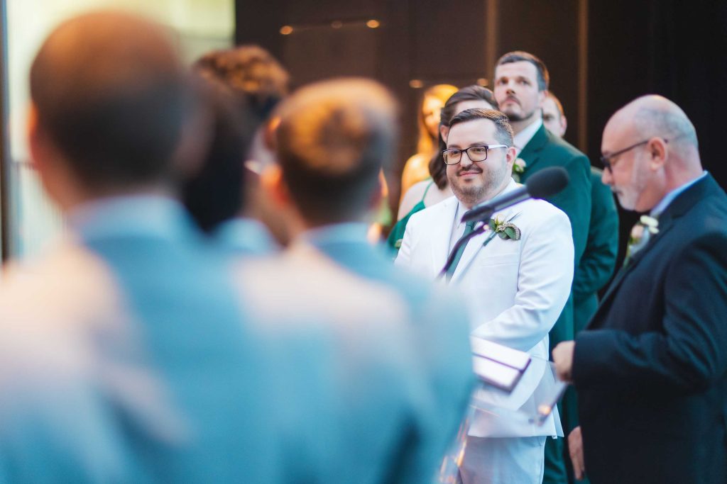 A man in a white suit stands at a wedding ceremony in Baltimore, smiling, while people in suits and formal attire look on.