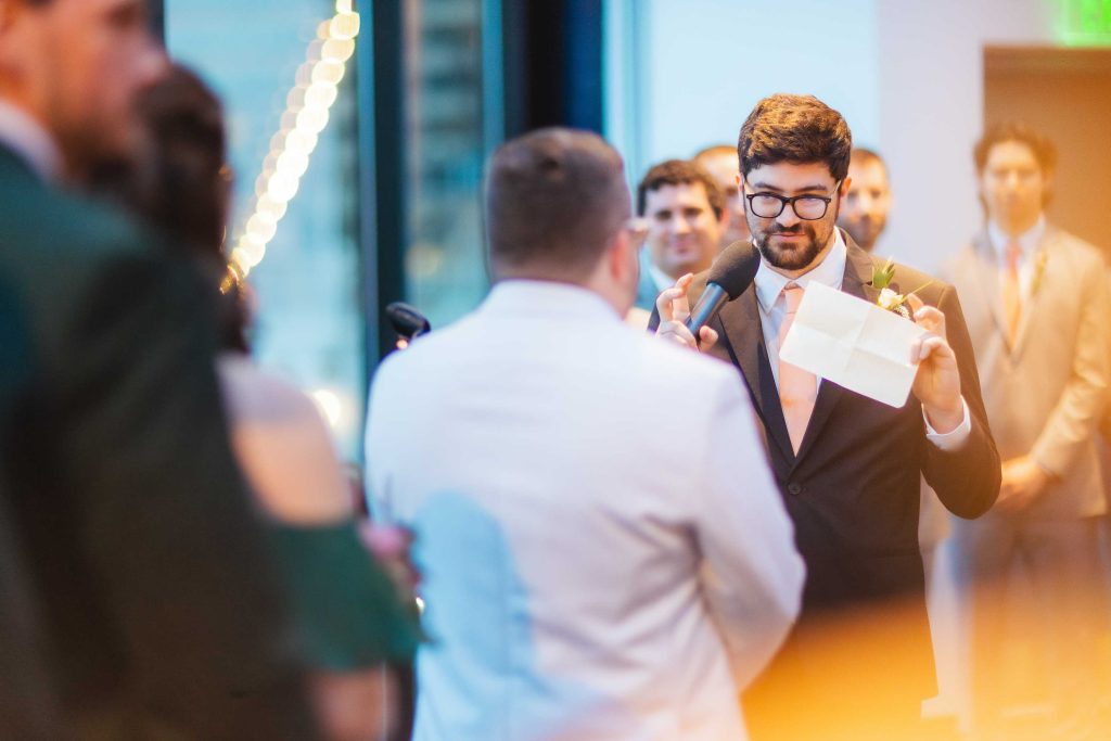 A man in a suit and glasses holds a microphone and reads from a piece of paper, addressing another man in a white jacket during a same sex ceremony at an indoor event with people in the background.