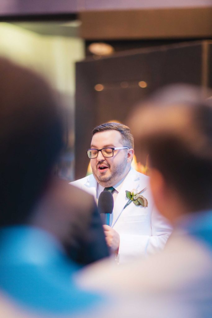 A man in a white suit and glasses speaks into a microphone at an event at the National Aquarium in Baltimore, with blurred people in the foreground.