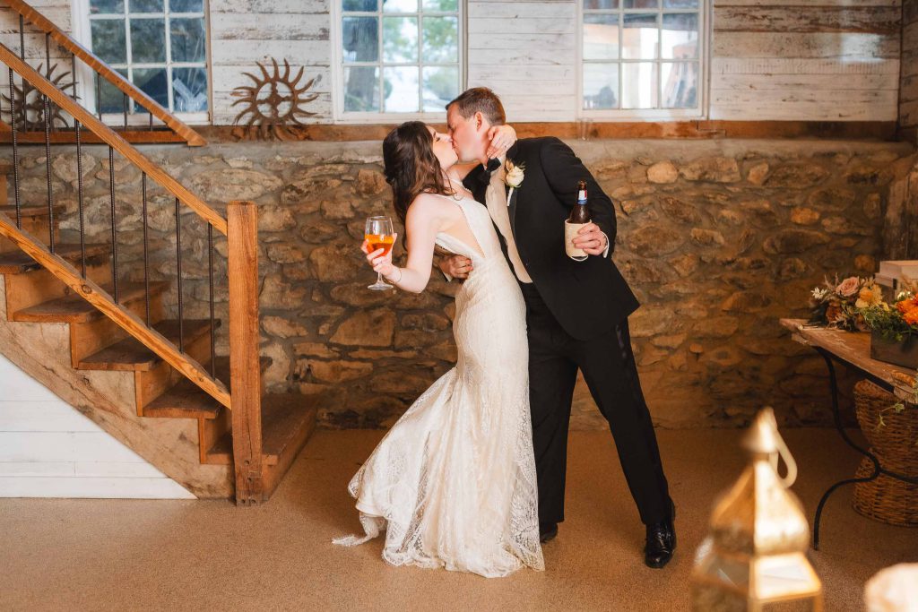 A bride and groom share a kiss while holding drinks in a rustic, stone-walled room with wooden stairs during their wedding reception at The Barns at Hamilton Station. Both are elegantly dressed, and the groom has a bottle in one hand.
