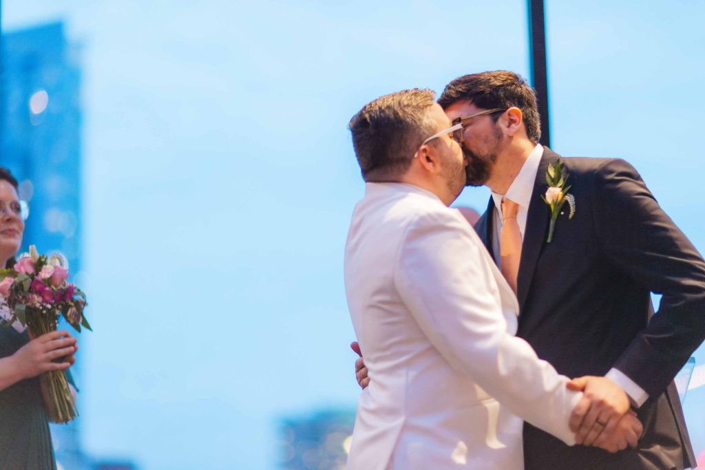 Two men, one in a white suit and the other in a dark suit, share a kiss at their Baltimore wedding ceremony, while a person holding a bouquet stands nearby.