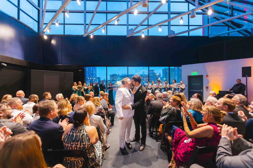Two grooms share a kiss at the front of their Baltimore wedding ceremony, surrounded by seated guests and breathtaking city skyline views through large windows.