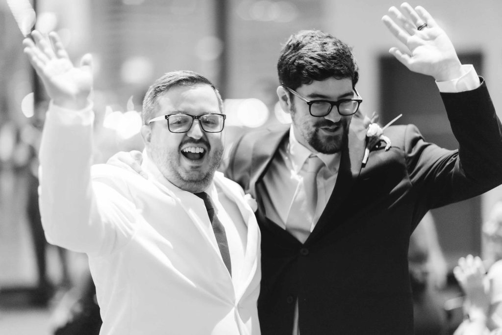Two men in suits, one in white and one in black, stand side by side smiling and waving with raised arms in a celebratory manner near Baltimore’s National Aquarium, celebrating their same sex union.