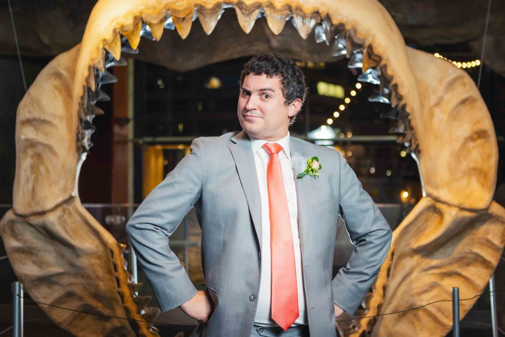 A man in a gray suit and orange tie stands with hands on hips in front of a large display of fossilized jaws with visible sharp teeth at the National Aquarium in Baltimore.