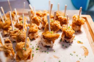 Several pieces of fried chicken on skewers sit atop small waffle squares, garnished with chopped chives and drizzled with syrup, on a parchment-lined tray—perfect for sharing after a visit to Baltimore’s National Aquarium.