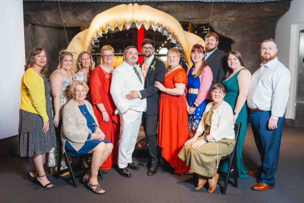 A group of people dressed formally, including two grooms at the center, pose for wedding portraits in front of a large shark jaw exhibit at the National Aquarium.