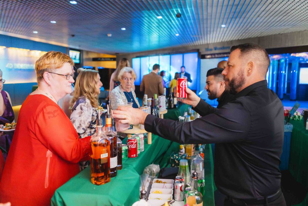 At a busy reception at the National Aquarium in Baltimore, a bartender serves drinks, including a can of Coca-Cola, to guests with bottles and mixers visible on the counter.