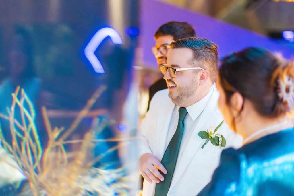 A man in a white suit with a boutonniere stands beside two people at a wedding reception, smiling and looking at an aquarium display with branches and neon lights in Baltimore.