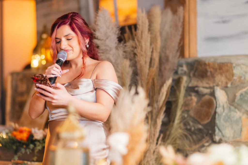 A woman with red hair is speaking into a microphone while holding a phone. She is wearing an off-shoulder dress and standing in front of a rustic, decorated background with pampas grass and warm lighting, perfect for a wedding reception at The Barns at Hamilton Station.