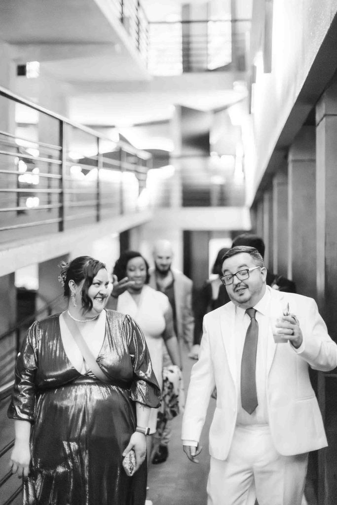 A group of people dressed formally walk down a hallway at a wedding reception. The person in front wears a shiny dress, and the person beside them, holding a drink, sports a light suit—capturing the joyful atmosphere in Baltimore.