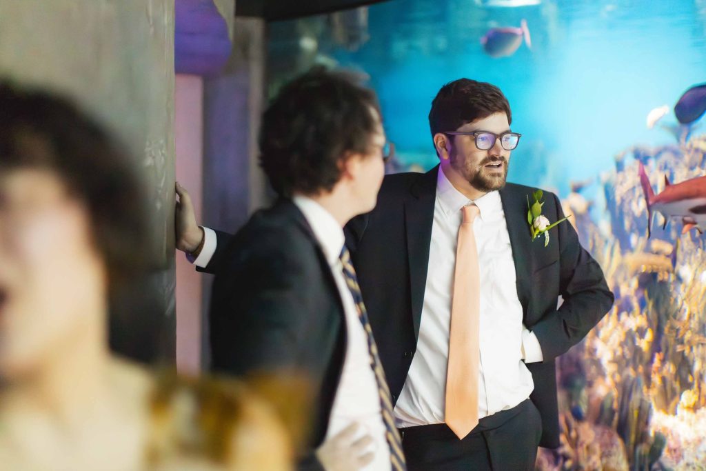 Two men in suits stand and talk near a large aquarium tank at the National Aquarium in Baltimore, with colorful fish swimming behind them. One man wears an orange tie and boutonniere, hinting at a wedding celebration.