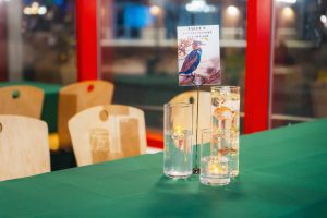 Three glass candle holders with floating candles and a bird-themed table sign add charming wedding details to a green tablecloth in a brightly lit room with wooden chairs, inspired by the vibrant atmosphere of the National Aquarium.