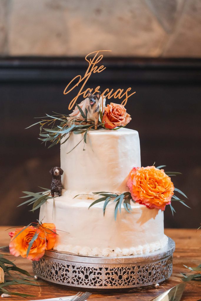 A two-tier white wedding cake with orange flowers and leaf decorations, topped with figurines of dogs and a sign that reads "The Jaquas," perfectly complements the charm of The Barns at Hamilton Station. Every detail exudes elegance on this special wedding day.