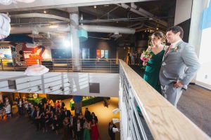 A well-dressed couple stands on an upper level balcony at the National Aquarium in Baltimore, overlooking a group of people gathered below at a wedding event space.