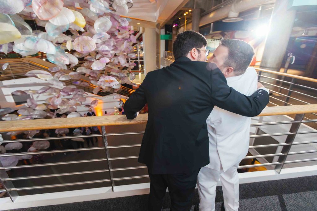 Two men in formal suits embrace by a railing at the National Aquarium in Baltimore, overlooking a busy reception area with hanging decorative elements.