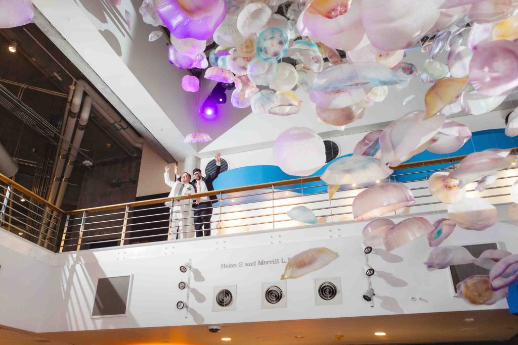 Three people stand on an upper balcony, waving during a reception, surrounded by a ceiling installation of translucent, jellyfish-like sculptures in the modern indoor space of Baltimore’s National Aquarium.