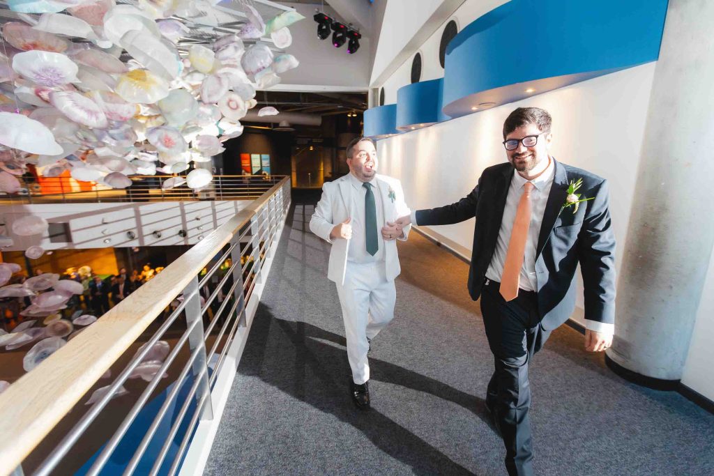 Two men in suits, one in white and one in black, walk together holding hands as newlyweds in a modern Baltimore building with artistic ceiling decorations, celebrating their wedding near the iconic National Aquarium.