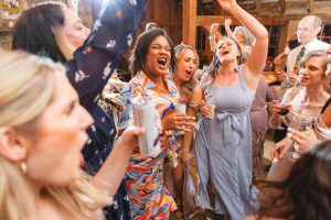 A group of people enthusiastically sing and dance at a lively wedding reception at The Barns at Hamilton Station, some holding drinks.