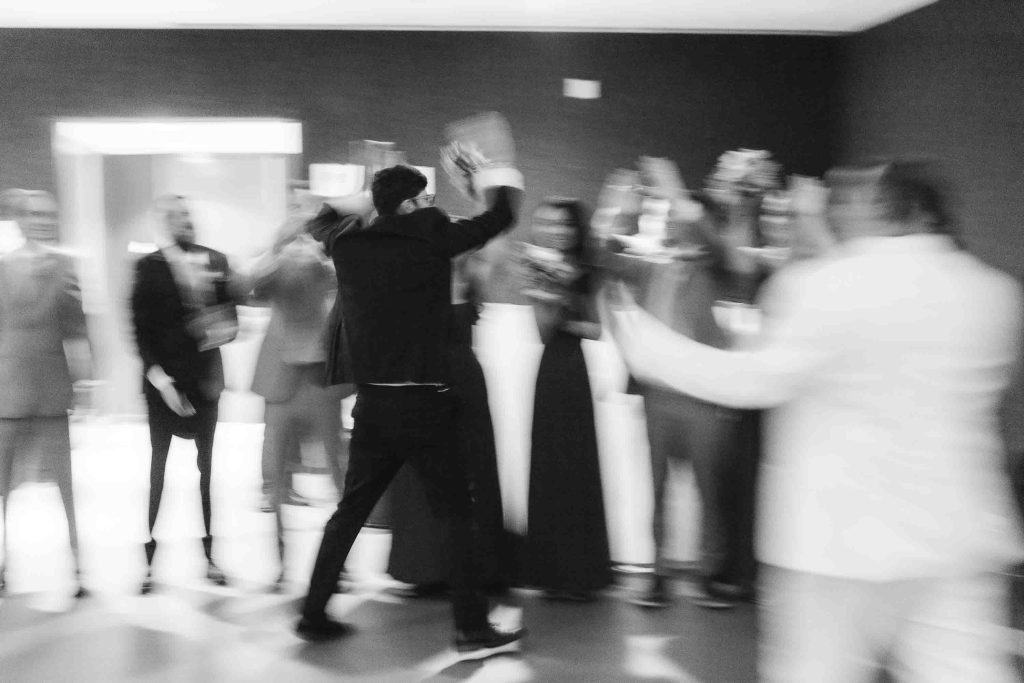 A person in a suit walks toward a group of people clapping and cheering at a wedding reception, all shown in motion blur in a monochrome setting.
