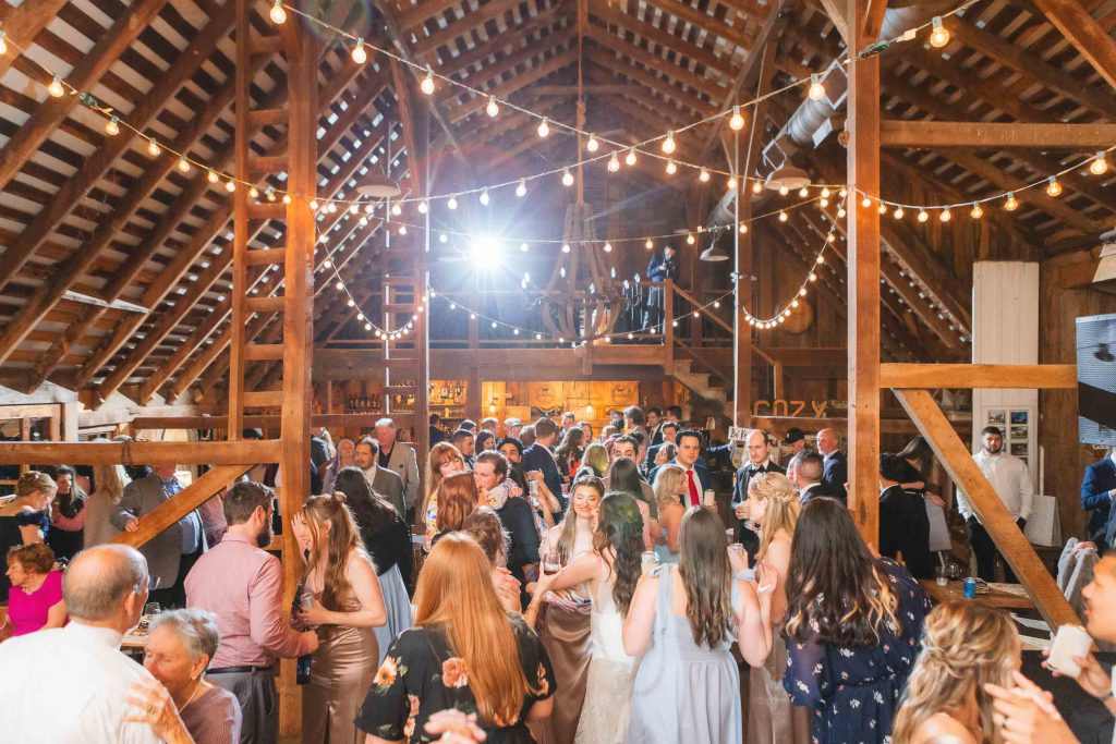 A large group of people dressed formally is gathered at The Barns at Hamilton Station, a wooden barn decorated with string lights, engaging in conversation and celebrating the wedding reception.