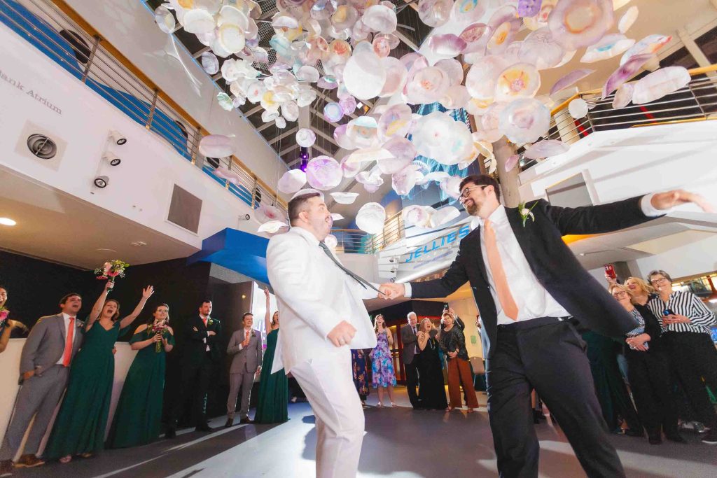 Two men in suits dance together at a Baltimore wedding in a large, well-lit room, with people watching and colorful decorations hanging from the ceiling.