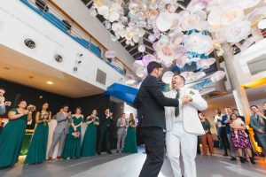 Two grooms, one in a black suit and one in a white suit, dance together at their Baltimore wedding reception while guests look on and floral decorations hang from the ceiling.