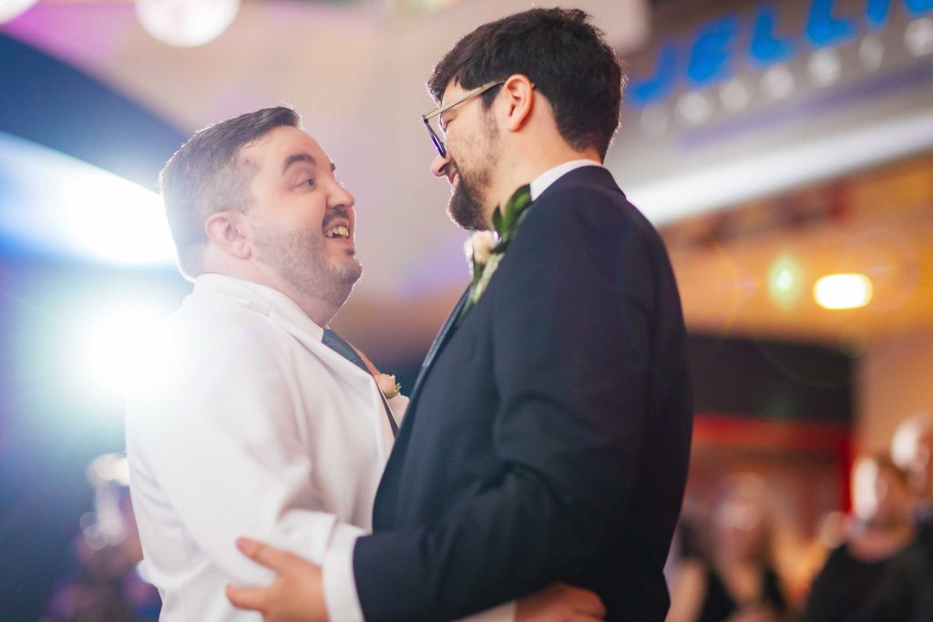Two men dressed in suits, one white and one black, smile and embrace while dancing together at their wedding indoors in Baltimore, with soft lighting glowing in the background.