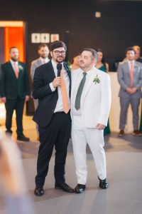 Two men stand together at a wedding reception, one in a black suit holding a microphone, the other in a white suit with a green tie; people in formal attire mingle in the background, evoking an elegant scene reminiscent of the National Aquarium's grand events.